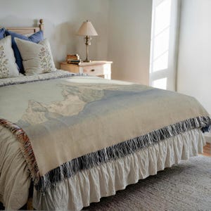 Puede incluir: Una manta beige con un diseño de paisaje montañoso, sobre una cama. La manta tiene un borde de flecos con detalles en azul y rojo. La cama tiene una falda de cama con volantes y almohadas decorativas.