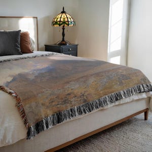 Op de afbeelding: Een geweven sprei met een landschapsontwerp in bruin-, beige- en blauwtinten is over een bed gedrapeerd. De deken heeft een franjerand met zwarte en witte draden. Een gebrandschilderde lamp staat op een zwart nachtkastje.