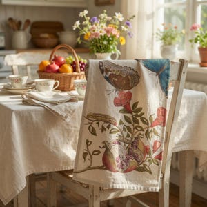 May include: A beige kitchen towel with a botanical print featuring butterflies, pomegranates, and flowers. The towel is draped over a white wooden chair in a sunlit kitchen setting. A basket of fruit and teacups are on the table.