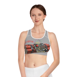 May include: Gray sports bra featuring a graphic of a cartoon bomb with red, black, and green details. The bra has a white trim and a racerback design. The bomb graphic includes a digital display.