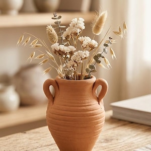 Peut inclure: Un vase en terre cuite avec deux anses, rempli de fleurs et de graminées séchées. Le vase présente un motif texturé et côtelé et repose sur une table en bois. L'arrangement comprend des fleurs blanches, des épis de blé et de l'eucalyptus.