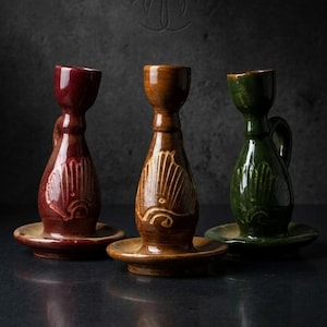 Puede incluir: Tres candeleros de cerámica en tonos rojo, marrón y verde. Cada uno tiene una forma única, un pequeño asa y un diseño decorativo. Se asientan sobre bases redondas a juego, creando una estética rústica.