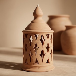 Peut inclure: Un photophore en terre cuite avec un motif ajouré décoratif. Le photophore a un sommet conique et une base avec les initiales "MT". Deux autres vases en terre cuite sont à l'arrière-plan. Le photophore mesure environ 15 cm de haut.