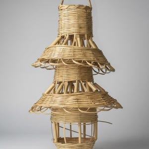 May include: A handcrafted, tiered wicker pendant light fixture. The natural-toned lamp features a cylindrical top with a handle, three flared tiers, and a bottom basket-like structure. The woven design creates a unique, decorative lighting element.