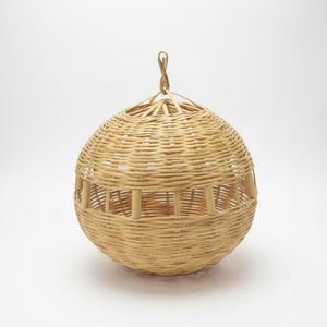 May include: A spherical, woven rattan basket with a natural finish. The basket has a circular opening at the top and a horizontal rectangular opening with vertical supports. A loop is attached at the top for hanging. The basket is designed for decorative or storage purposes.