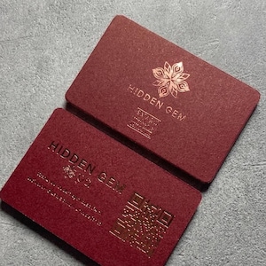 Puede incluir: Dos tarjetas de visita burdeos con texto dorado y un diseño floral. Las tarjetas dicen "HIDDEN GEM" e incluyen un código QR. Las tarjetas son rectangulares con esquinas redondeadas y están apiladas sobre una superficie gris.