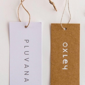 May include: Two rectangular product tags. One is white with the word "PLUVANA" printed vertically in gray. The other is brown with the word "OXLEY" printed vertically in white. Both tags have a hole at the top with a string attached.