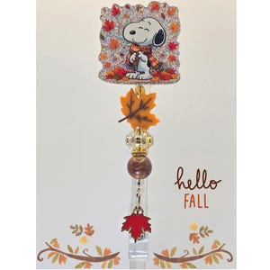 May include: A badge reel featuring a square glittery topper with a Snoopy illustration wearing a plaid scarf amidst autumn leaves. Below the topper are orange leaf, clear faceted, and brown beads, ending with a red maple leaf charm.