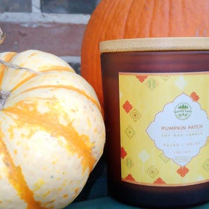 Puede incluir: Una vela decorativa marrón con tapa de madera, con la etiqueta "Pumpkin Patch Soy Wax Candle". Se encuentra junto a una calabaza blanca y naranja y una gran calabaza naranja. Decoración otoñal.