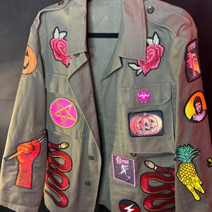 May include: Olive green jacket adorned with various patches, including a pink pentagram, a red snake, a jack-o'-lantern, and a pineapple. The jacket also features patches with text and images of a rose and a lightning bolt.