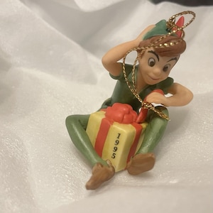 May include: A Peter Pan figurine ornament wearing a green tunic and cap with a red feather. The character is sitting and holding a yellow gift box with red ribbons and the year 1995 printed on the side. A gold hanging cord is attached to the top.