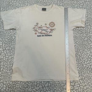 Puede incluir: Camiseta color crema con un diseño de mapa bordado en marrón y rojo, con el texto "Ilha Da Madeira" en azul marino. El mapa incluye símbolos y nombres de lugares. La camiseta tiene mangas cortas y cuello redondo.