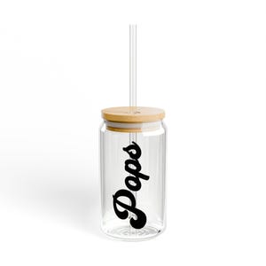 Puede incluir: Un vaso de vidrio transparente con tapa de bambú y pajita transparente. La palabra "Pops" está escrita en negro en el lateral del vaso. El vaso está diseñado para contener bebidas.