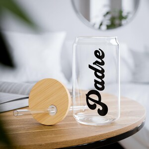 May include: A clear glass can-shaped drinking glass with the word "Padre" in black script. The glass has a bamboo lid and a clear straw. The glass sits on a wooden table.