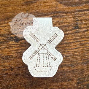May include: A white, windmill-shaped bookmark with brown stitched details. The windmill design features blades, a central structure, and a base. The bookmark has a tab at the top. The text "Kievel Kreations - Handcrafted with Love" is visible.