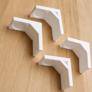 May include: Four white L-shaped corner protectors with a smooth matte finish. Each piece features a recessed circular indentation on both inner faces, designed to fit snugly against the edges of a frame or box to provide structural support.