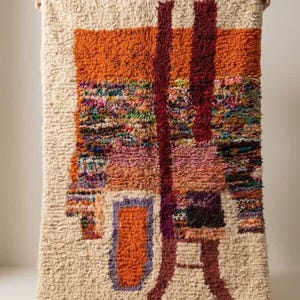 Peut inclure: Un tapis rectangulaire et texturé avec un motif abstrait. Le tapis présente un fond crème avec des formes géométriques orange, bordeaux et multicolores. Le tapis a une frange le long du bord inférieur et est tenu par des mains invisibles.