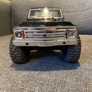 May include: Black toy truck with a chrome grill and headlights. The truck has large, textured tires and a clear windshield. The front of the truck is in focus, showing the details of the bumper and the Chevrolet emblem.