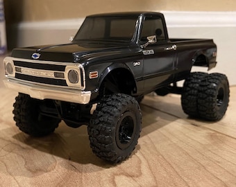May include: A black Chevrolet pickup truck model with a chrome front bumper, grille, and side mirrors. The vehicle features large, textured off-road tires, a dark tinted windshield, and the text SCX10 and DAD printed on the side of the body panel.