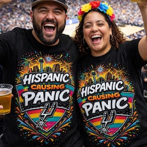 May include: Two people wearing black t-shirts with the text "HISPANIC CAUSING PANIC" and a colorful graphic. One person wears a Spurs baseball cap and the other a floral crown. Both are smiling and holding drinks.