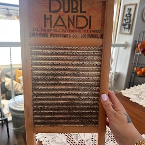 Op de afbeelding: Vintage houten wasbord met de tekst "DUBL HANDI" en "COLUMBUS WASHBOARD CO, COLUMBUS, A". Het wasbord heeft een gegolfd metalen schrobvlak en een houten frame. Het wasbord is bruin en beige.