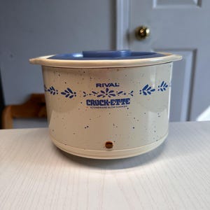 May include: Cream-coloured Rival Crock-ette stoneware slow cooker with blue floral accents and a blue lid. The slow cooker has a speckled design and the brand name is visible on the front.