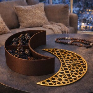 May include: A brown, crescent-shaped serving dish filled with dried fruit, next to a gold-colored, crescent-shaped decorative piece with a geometric pattern. A lit lantern and prayer beads are also on the table. The scene is set on a dark table with a sofa in the background.