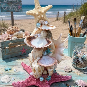 May include: A decorative seashell sculpture with a pink starfish base, decorated with various shells, pearls, and small blue starfish. The sculpture is on a weathered, light blue wooden surface, with a beach scene in the background.