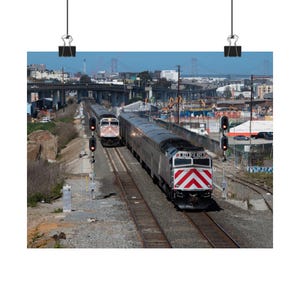 Puede incluir: Dos trenes de cercanías que viajan por vías paralelas, vistos desde arriba. El tren delantero tiene un diseño de chevrón rojo y blanco. El fondo incluye un puente, edificios y equipos de construcción.