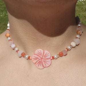 May include: A beaded choker necklace featuring a central light pink carved flower pendant. The necklace is composed of alternating white pearls, translucent light pink stone beads, and small orange spherical beads arranged in a symmetrical pattern.