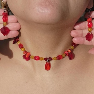 May include: A necklace and matching pair of drop earrings featuring red and yellow beads. The necklace has a central red teardrop pendant, while the earrings consist of red and yellow beads with a red flower-shaped charm at the base.