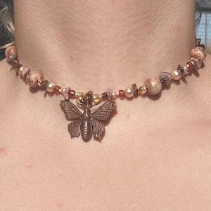 May include: A beaded choker necklace featuring a central bronze-toned butterfly pendant. The necklace is composed of a variety of round, marbled, and small metallic beads in shades of brown, tan, cream, and pink, strung together in a repeating pattern.