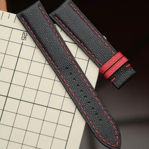May include: A black textured watch strap with red stitching and two red leather keepers, positioned next to a grid ruler showing a 25 mm measurement mark. The strap features a pointed end and a series of punched holes for adjustment.