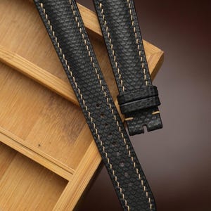 Puede incluir: Correas de reloj negras con cuero texturizado y costuras beige. Las correas se muestran en una bandeja de madera. Las correas de reloj tienen un diseño clásico y son adecuadas para varios estilos de reloj.