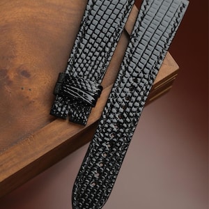 May include: Two black watch straps with a textured, reptile-like pattern. The straps are made of a shiny material and are displayed on a wooden surface. One strap has a buckle, and the other is a long, tapered piece.