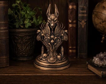 May include: A bronze-colored Anubis bust statue with intricate details, including a jackal head, ornate collar, and skull accents. The statue is displayed on a wooden shelf with books and a potted plant in the background.