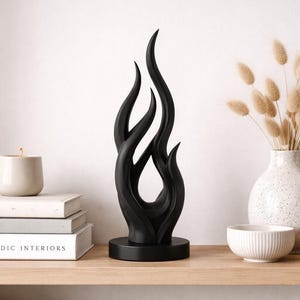 Könnte beinhalten: Schwarze, abstrakte Flammens Skulptur mit glatter Oberfläche, auf einem kreisförmigen Sockel. Das dekorative Objekt steht auf einem Holzregal mit Büchern und einer Vase. Modernes Wohnaccessoire.