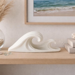 May include: A white ceramic sculpture shaped like a stylised ocean wave with two crests. The sculpture has a smooth, matte finish and a curved, flowing form that mimics the motion of water. It is positioned horizontally on a flat surface.