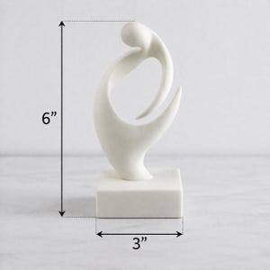 May include: A white abstract sculpture featuring a smooth, curved form mounted on a square base. The sculpture measures 6 inches in height and 3 inches in width, as indicated by the black dimension lines and arrows visible in the image.