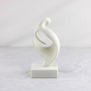 May include: A white abstract sculpture featuring a smooth, curved, and fluid form that rises from a solid square base. The object has a matte finish and consists of interconnected, sweeping lines that create an open, hollow center.