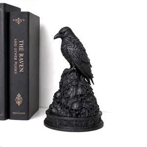 May include: A black raven figurine perched atop a mound of skulls and tangled vines, set on a circular base with architectural detailing. Next to it are two black books, one titled Edgar Allan Poe and the other The Raven and Other Poems.
