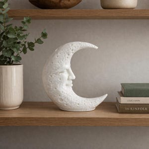Könnte beinhalten: Eine weiße Mondsichel-Skulptur mit einer texturierten, kraterartigen Oberfläche und einem menschlichen Gesichtsprofil. Der Mond steht auf einem Holzregal neben einem Stapel aus zwei Büchern und einer hellen Keramikvase mit einer grünen Pflanze.