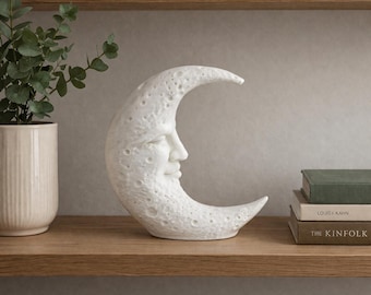 May include: A white crescent moon sculpture featuring a textured, cratered surface and a human-like face profile. The moon is positioned on a wooden shelf next to a stack of two books and a light-colored ceramic vase containing a green plant.