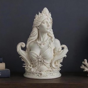 May include: A white sculpture of a figure with long flowing hair, wearing a crown and a necklace featuring a shell pendant. The figure is adorned with intricate details including seashells and coral motifs at the base and sides of the bust.