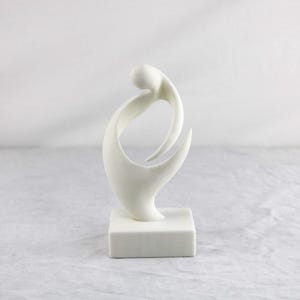 May include: A white abstract sculpture featuring a smooth, curved, organic form that loops upward from a solid square base. The piece has a matte finish and consists of continuous, flowing lines that create an open, negative space in the center.