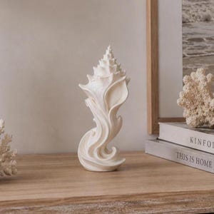 May include: A cream-coloured decorative sculpture shaped like a tall, spiralling seashell with a pointed top and a flowing, wave-like base. The object features a smooth, matte finish and intricate, curved ridges that mimic the natural form of a conch shell.