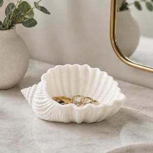 May include: A white seashell-shaped trinket dish with a spiral design, holding gold rings. The dish sits on a marble surface, next to a white vase with greenery and a gold-framed mirror.