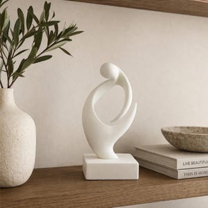 May include: A white abstract sculpture featuring smooth, curved lines that form an interlocking shape, mounted on a solid square base. The sculpture is positioned on a wooden surface next to a textured vase, a stone bowl, and stacked books.