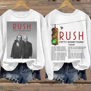 May include: White long-sleeved sweatshirt featuring a graphic of the band Rush. The front of the shirt displays the band's name, tour title, and a photo of the band members. The back of the shirt has a traffic light graphic and tour dates.