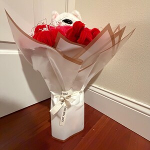 Könnte beinhalten: Ein Strauß aus roten Plüschblumen und einer weißen Stoffkatze, verpackt in weißem Papier mit Goldkante. Der Strauß ist mit einem Band mit der Aufschrift "Just For You" gebunden.